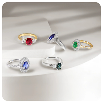 Five rings with colored gemstones and clear accent stones, displayed on a white geometric surface. The rings feature yellow gold and silver bands with oval, marquise, and round-cut gemstones in red, blue, and green tones.