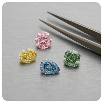 Four loose colored gemstones in pink, yellow, blue, and green are arranged on a brushed metal surface beside a pair of metal tweezers.