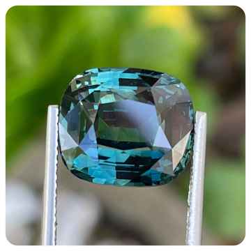 Faceted teal sapphire gemstone held between two tweezers with a blurred green background