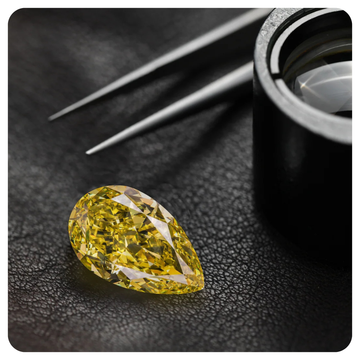A yellow pear-cut diamond gemstone displayed on a textured black surface, with tweezers and a loupe microscope in the background.