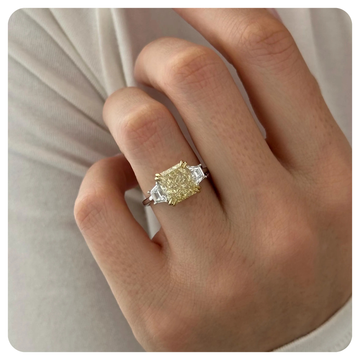 A close-up of a hand wearing a yellow diamond ring with silver-colored side stones on a neutral beige background.